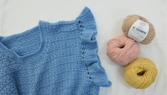 Maglia azzurra lavorata a mano in cashmere rigenerato con dettaglio volant sulla manica, accanto a gomitoli Re-Cashmere Bettaknit in tonalità beige, rosa e giallo su sfondo chiaro.