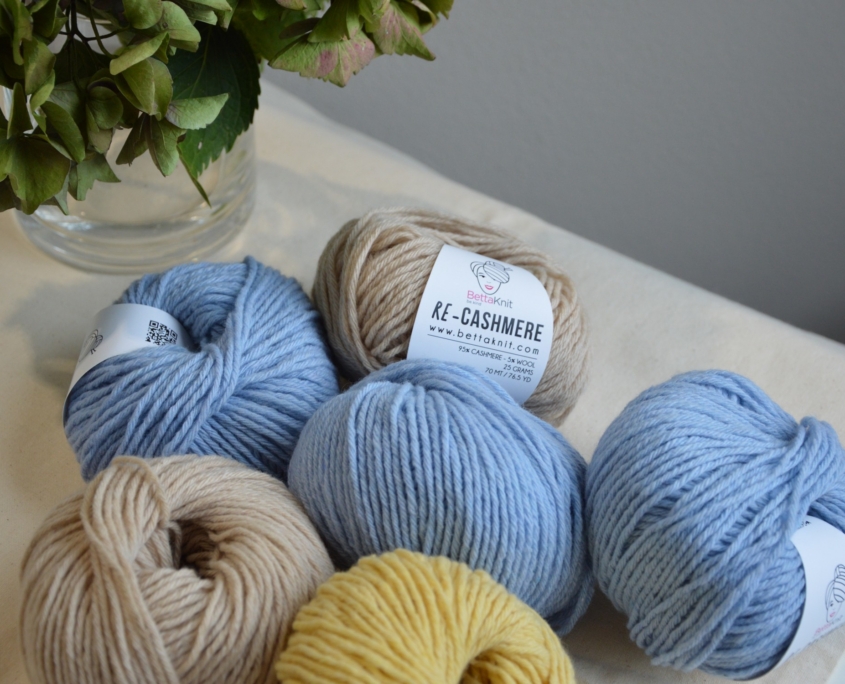 Gomitoli di cashmere rigenerato Re-Cashmere Bettaknit nei colori azzurro, beige e giallo disposti su tessuto chiaro accanto a un vaso con fiori.