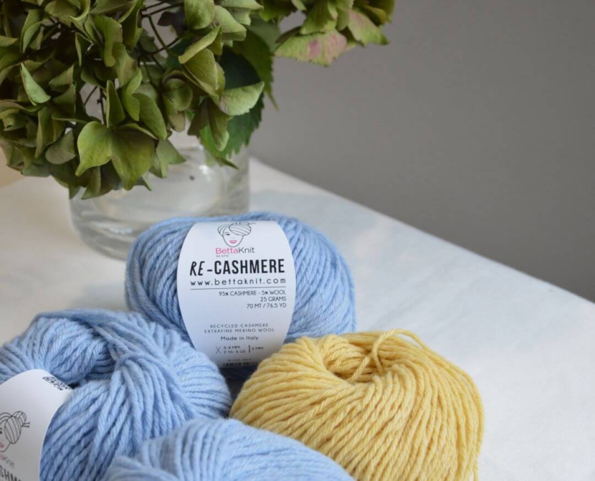 Gomitoli di cashmere rigenerato Re-Cashmere Bettaknit nei colori azzurro, beige e giallo disposti su tessuto chiaro accanto a un vaso con fiori.